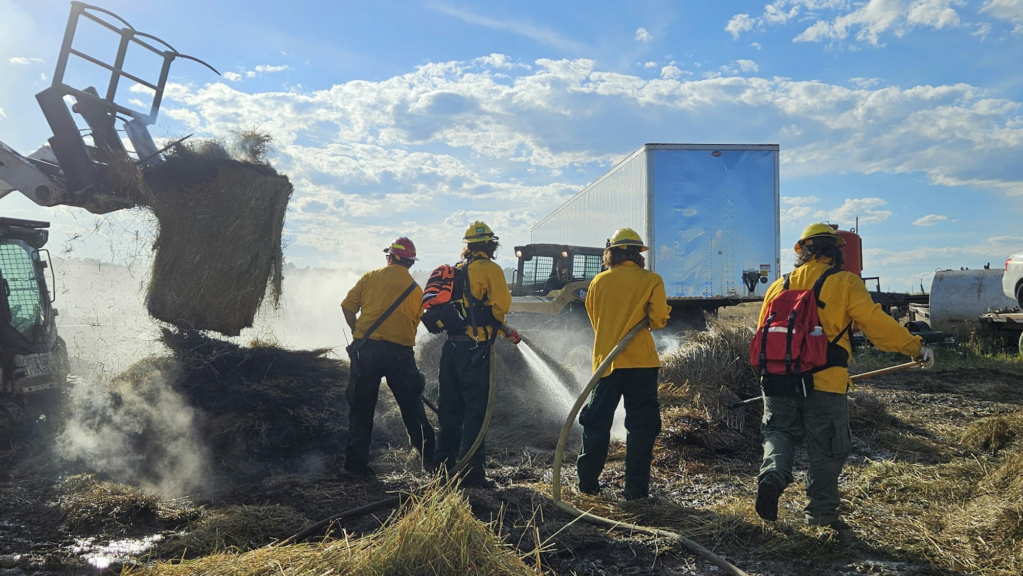Firefighters and a skid steer working to extinguish a fire near a truck.