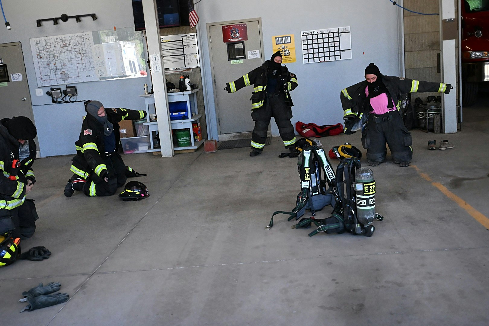 Firefighters gear up in a station, preparing equipment, with maps and gear visible on the walls.