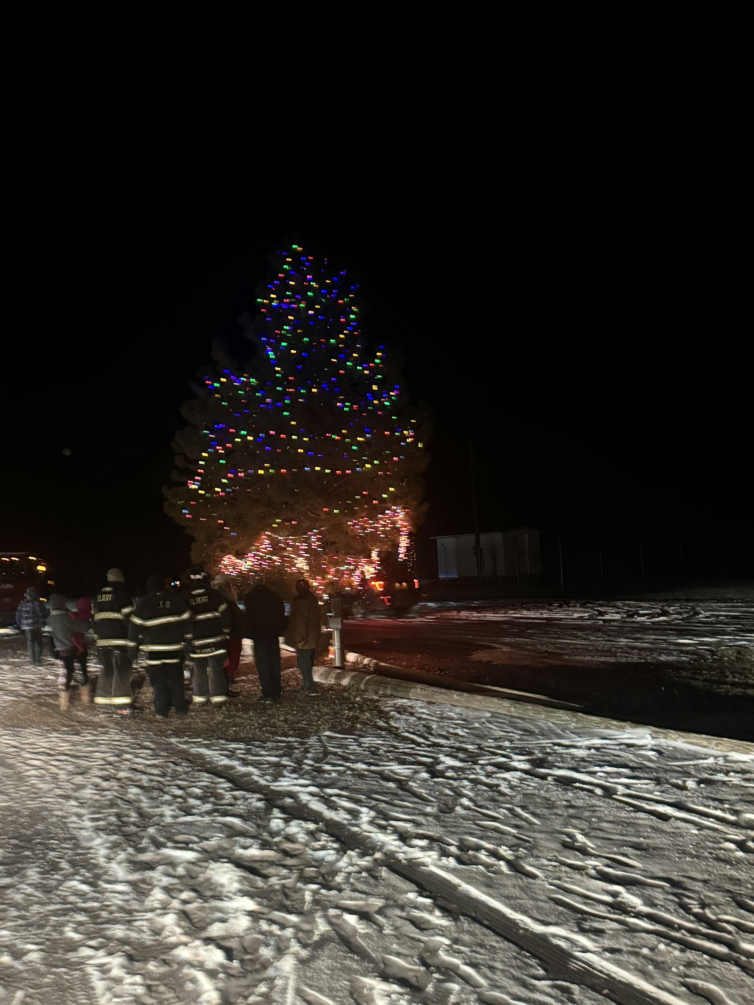 A Christmas tree decorated with colorful lights outside, with people, including firefighters, on a snowy night.