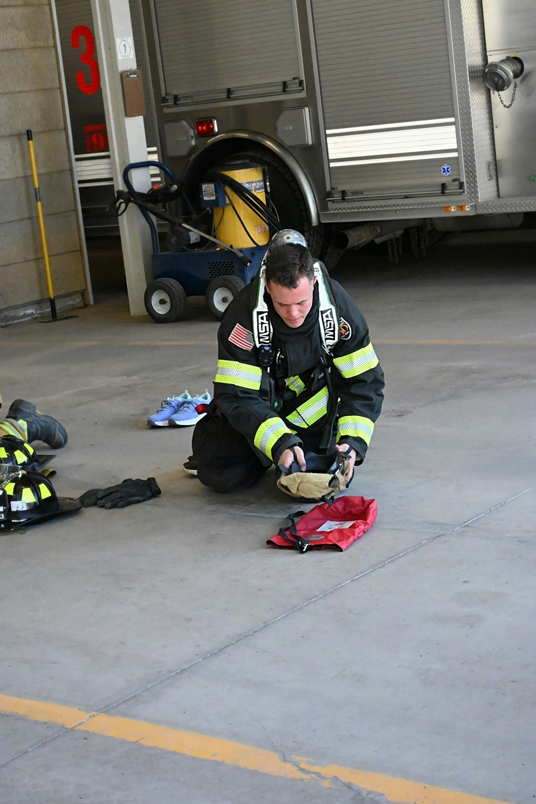 A firefighter kneels and prepares equipment in a fire station, with helmets and gloves nearby.