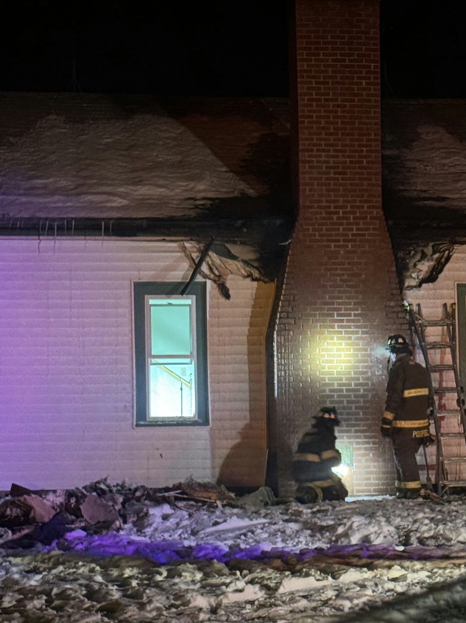 Firefighters inspecting a house with exterior fire damage at night. Snow and debris are visible on the ground.