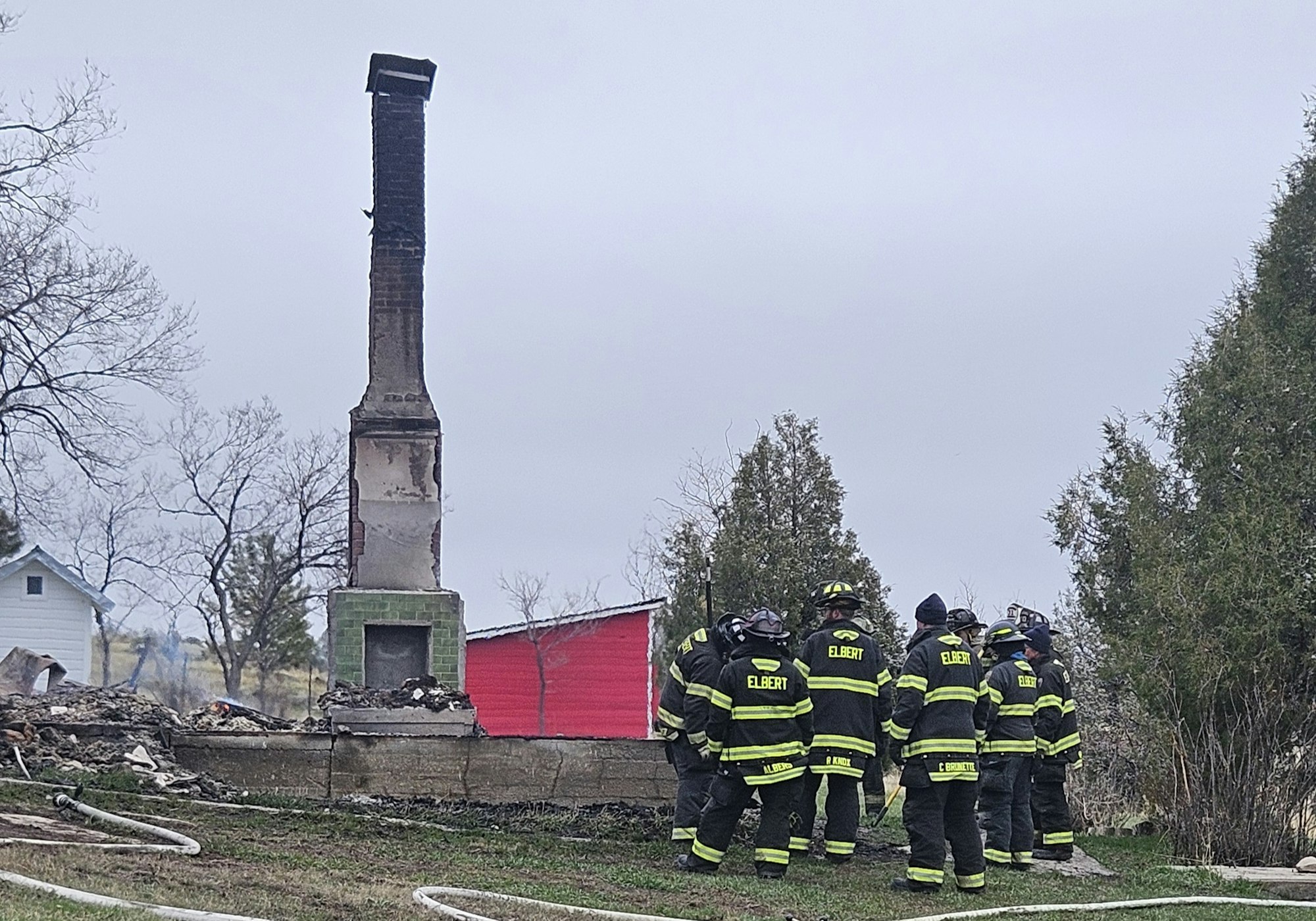 Firefighters near a chimney, the remains of a house after a fire, with a red shed in the background.
