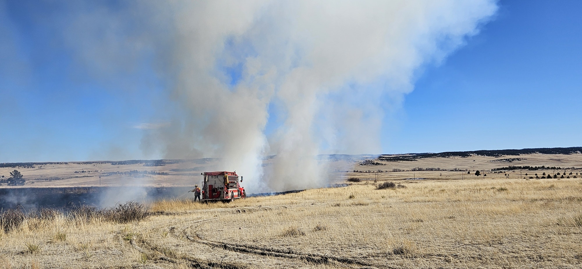 A fire truck and firefighter battle a grass fire in a rural area with smoke billowing into the sky.