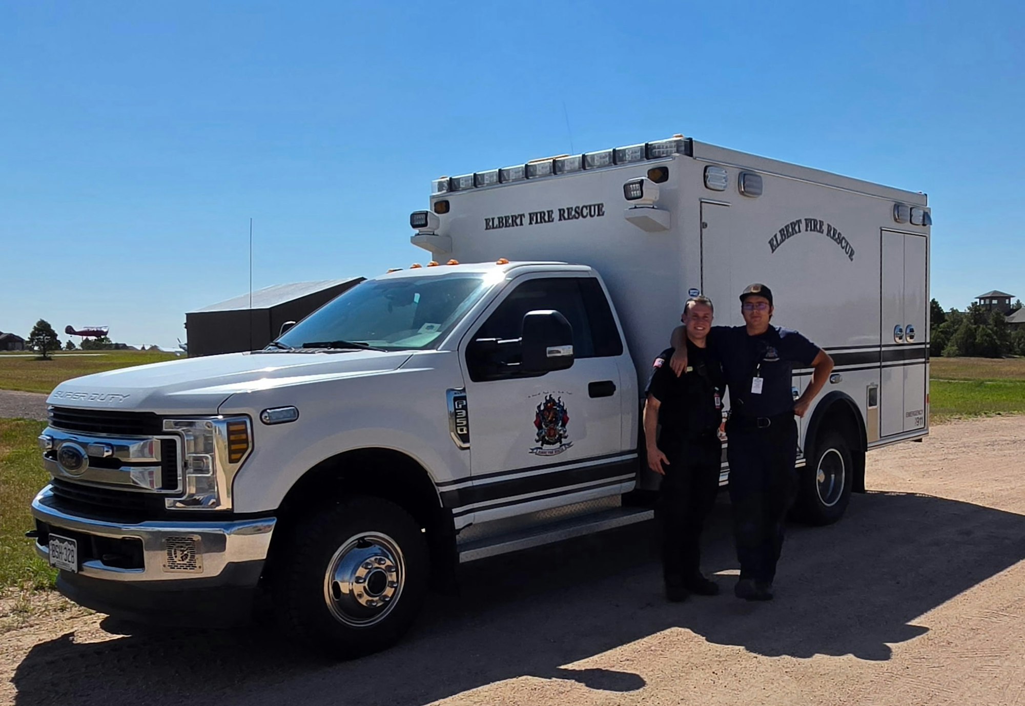 The image features two firefighters posing beside a white Elbert Fire Rescue truck in a sunny outdoor setting.