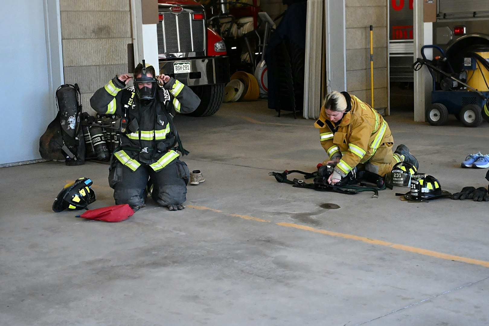 Two firefighters in gear, one adjusting a mask, the other handling equipment, with a fire truck and helmets nearby.