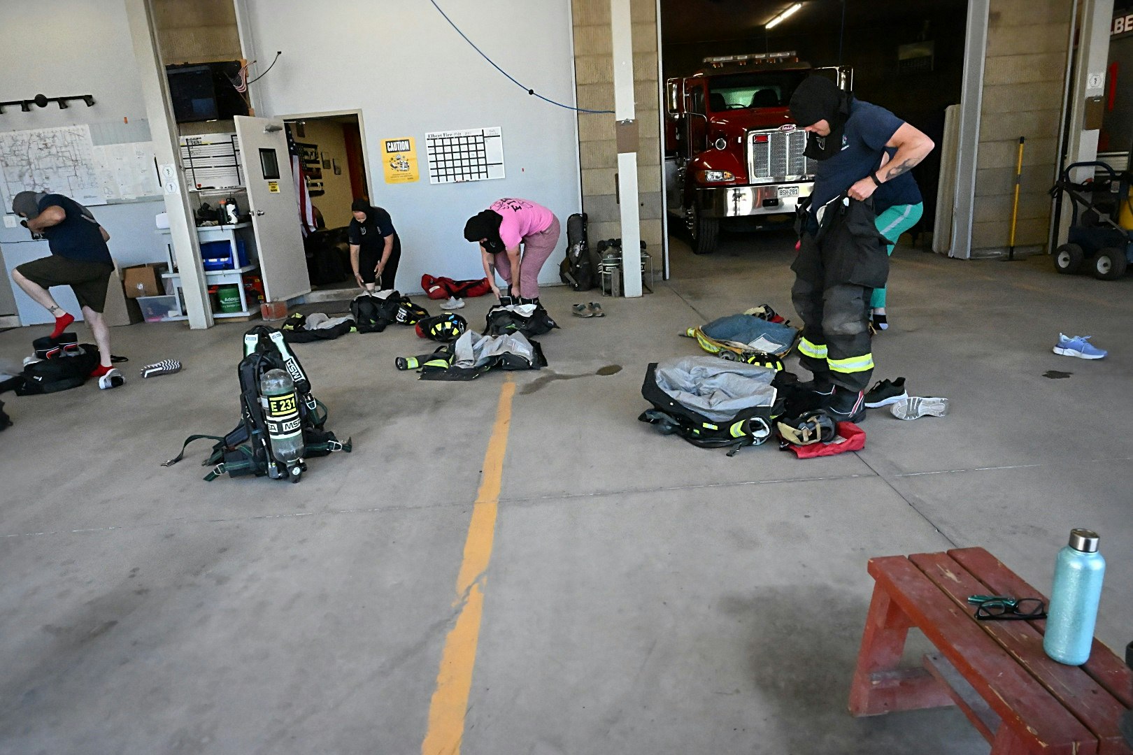 People in a fire station put on firefighting gear beside a fire truck.