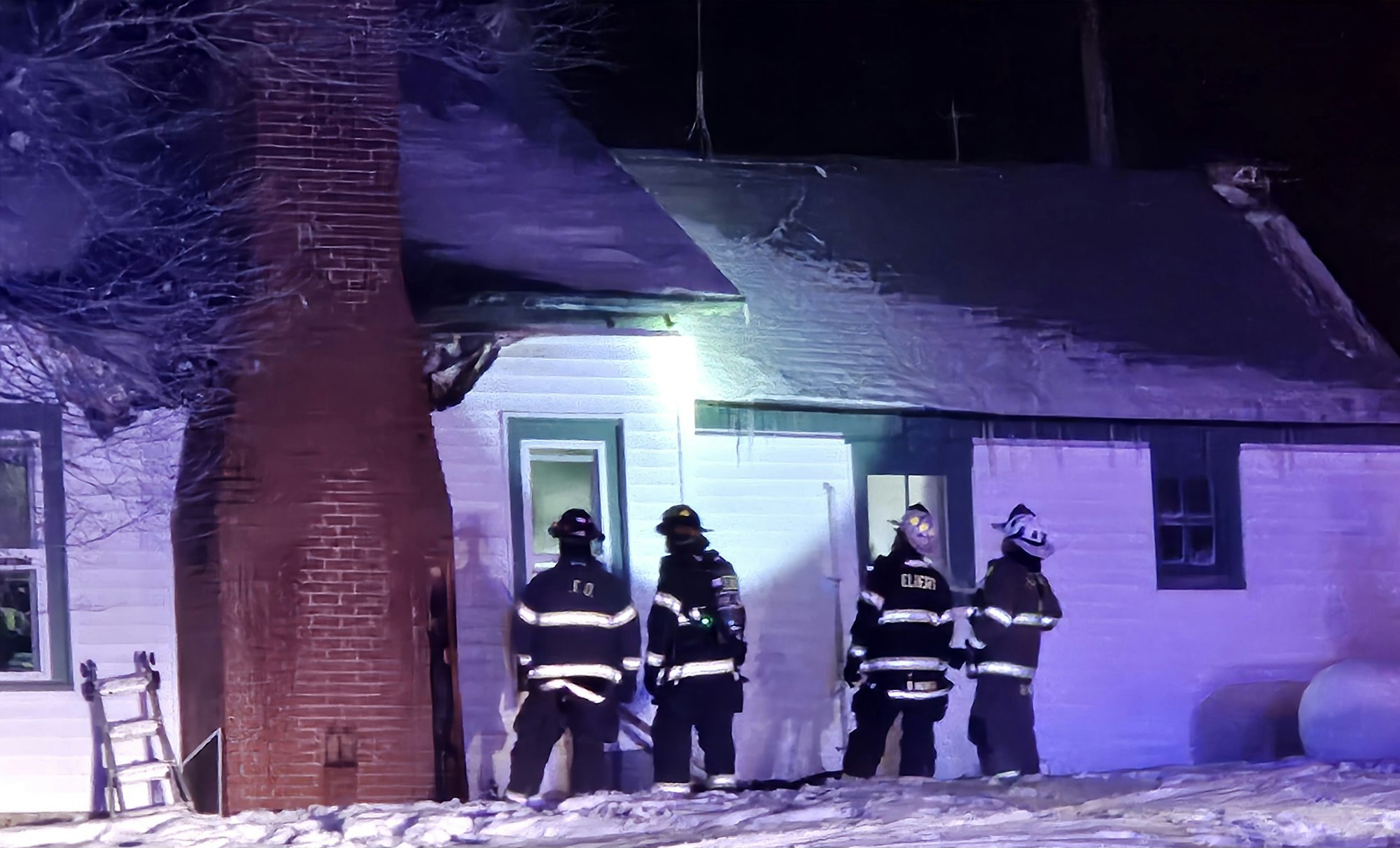 Firefighters in gear standing outside a snowy house with a brick chimney at night.