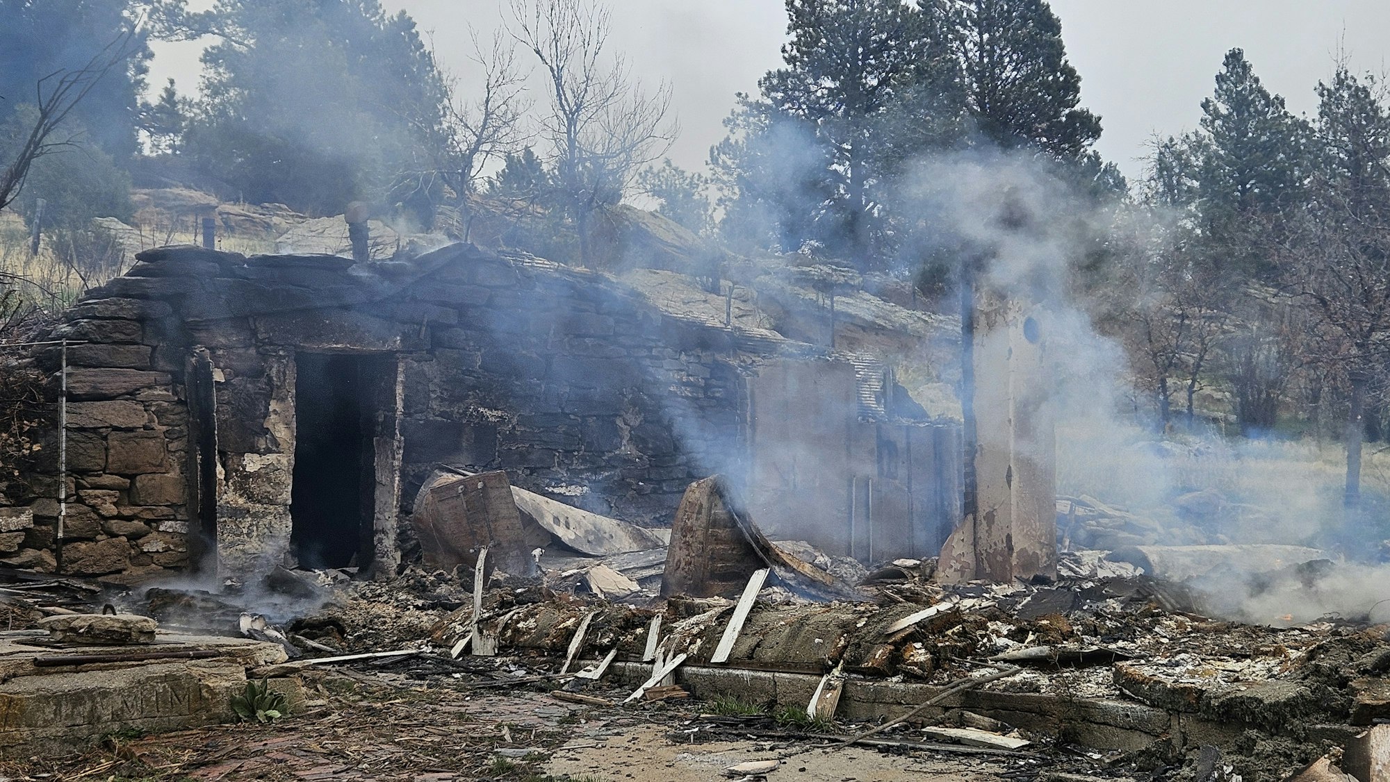 Burnt remnants of a building with smoke, amidst trees.