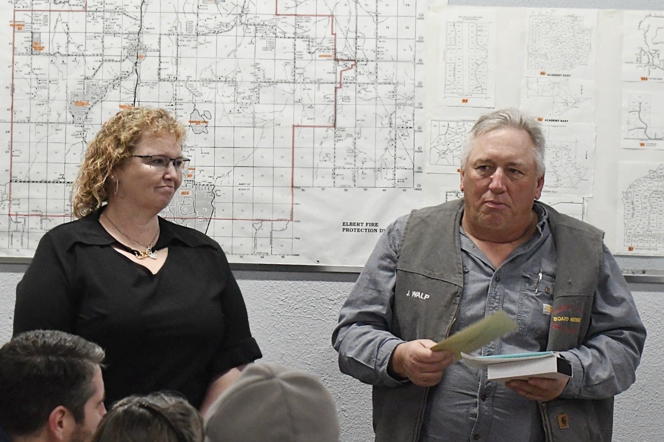 Two people stand in front of maps, one holding papers.