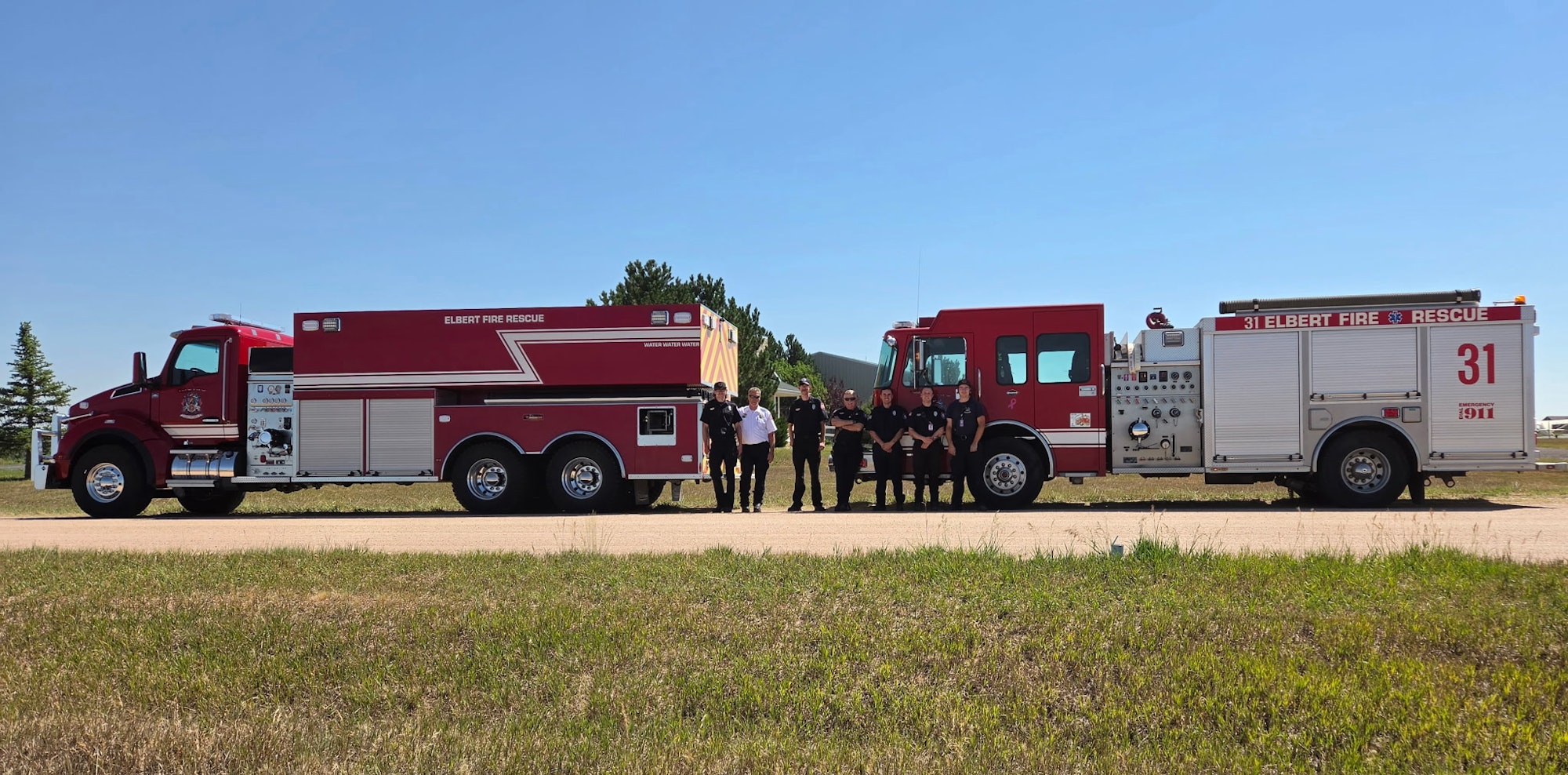 A fire rescue team stands in front of two red fire trucks on a sunny day, surrounded by grass and trees.