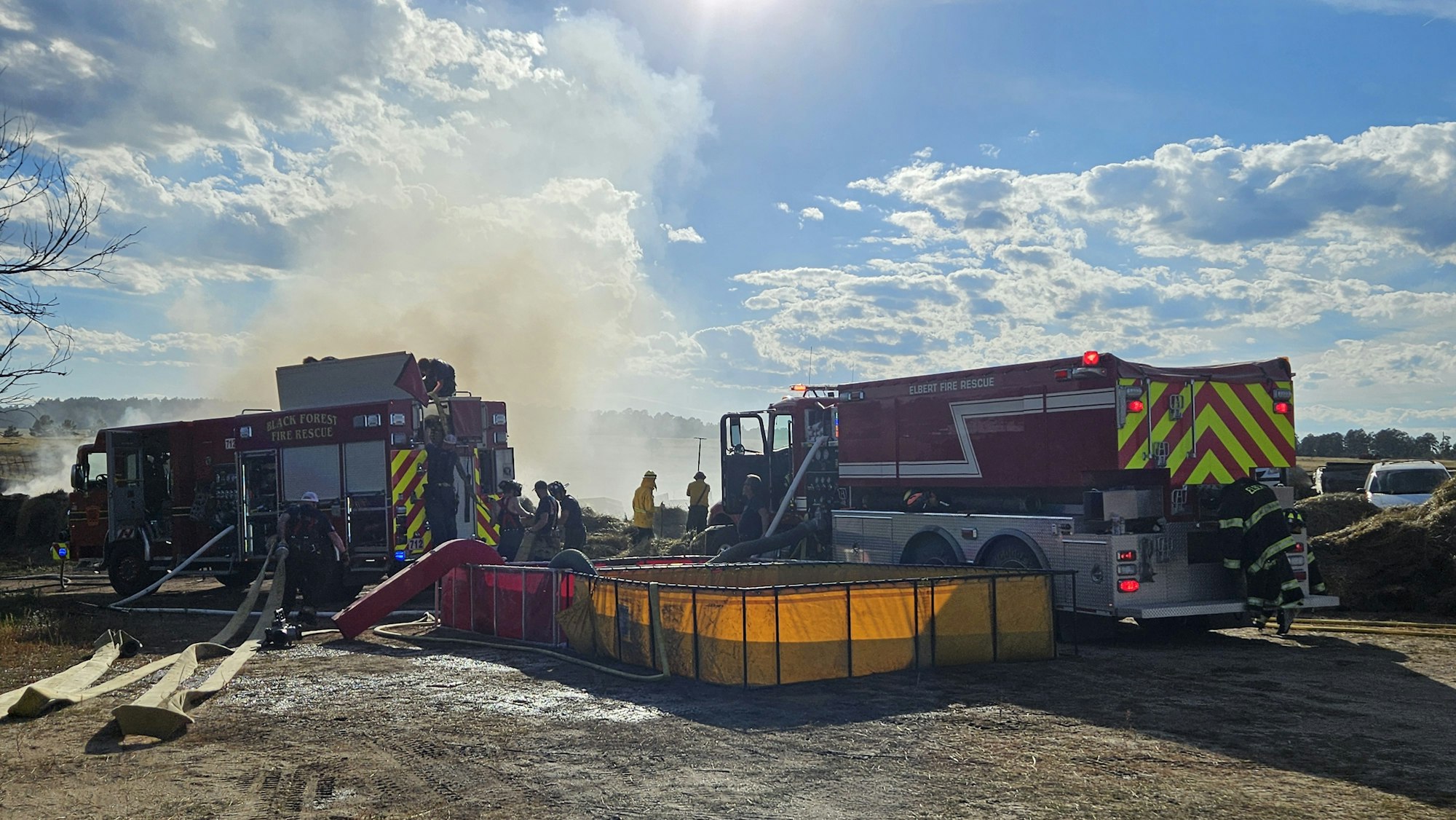Firefighters at a scene with fire trucks and equipment, possibly tackling a fire. Smoke and clear skies visible.