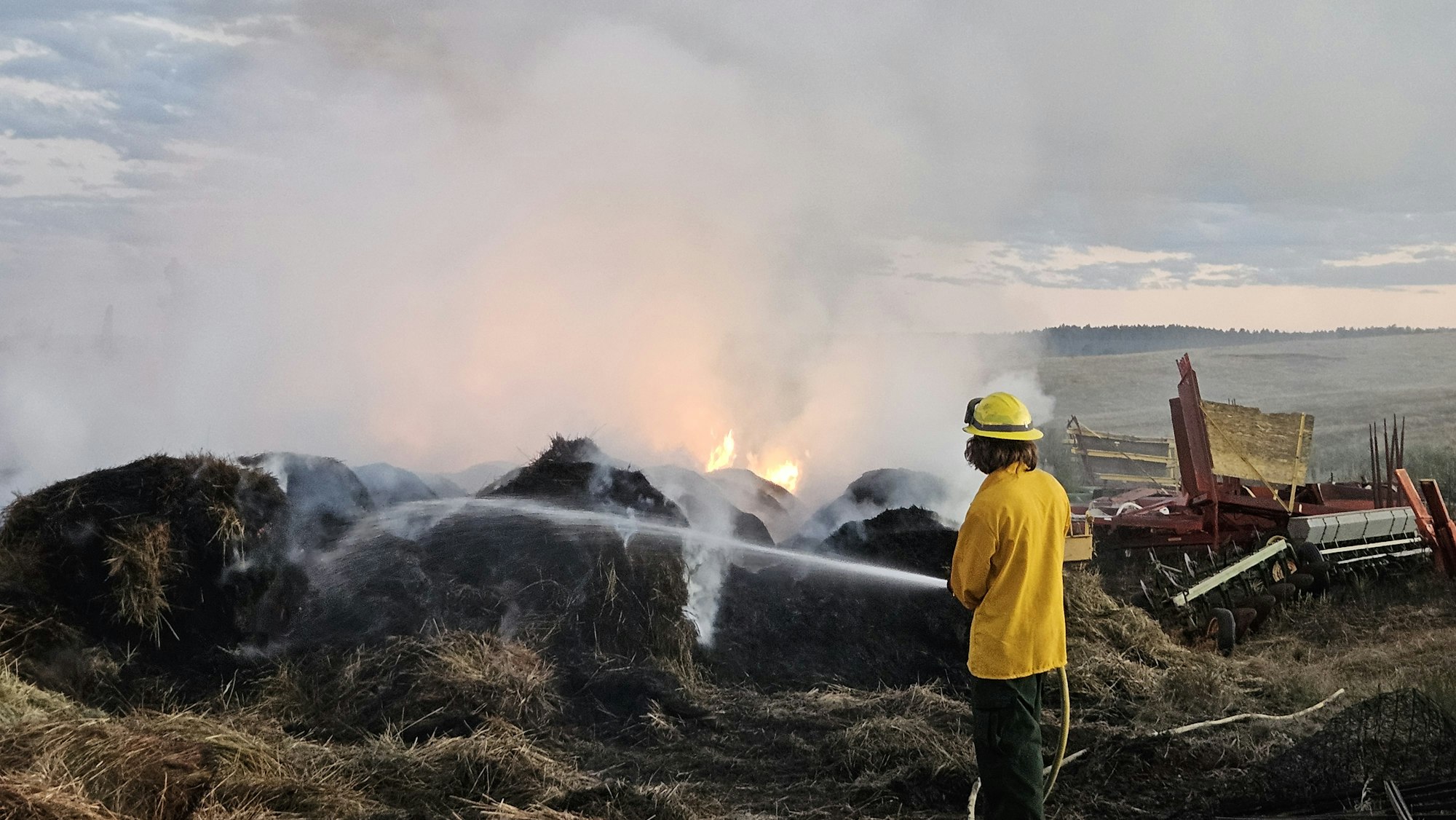 A firefighter is extinguishing a hay fire with a hose near some farm equipment. Smoke fills the air.