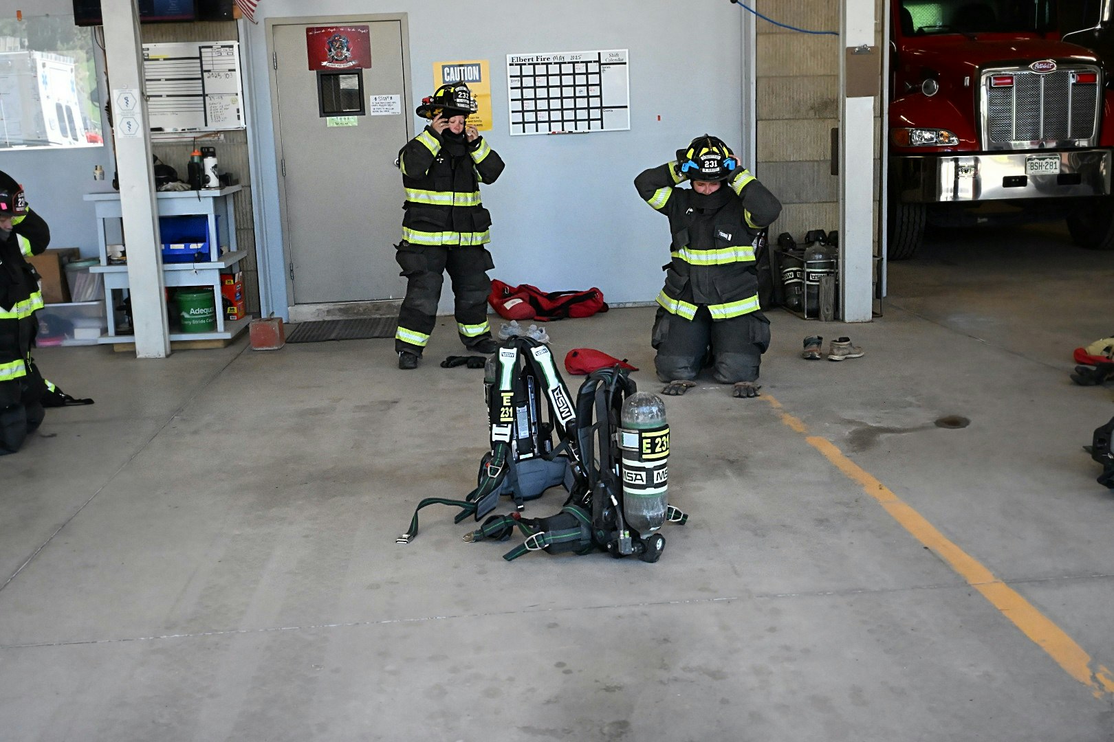 Firefighters in gear adjusting helmets indoors, with equipment and a fire truck nearby.