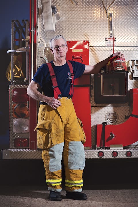 A firefighter in gear standing beside a fire truck, holding a helmet.