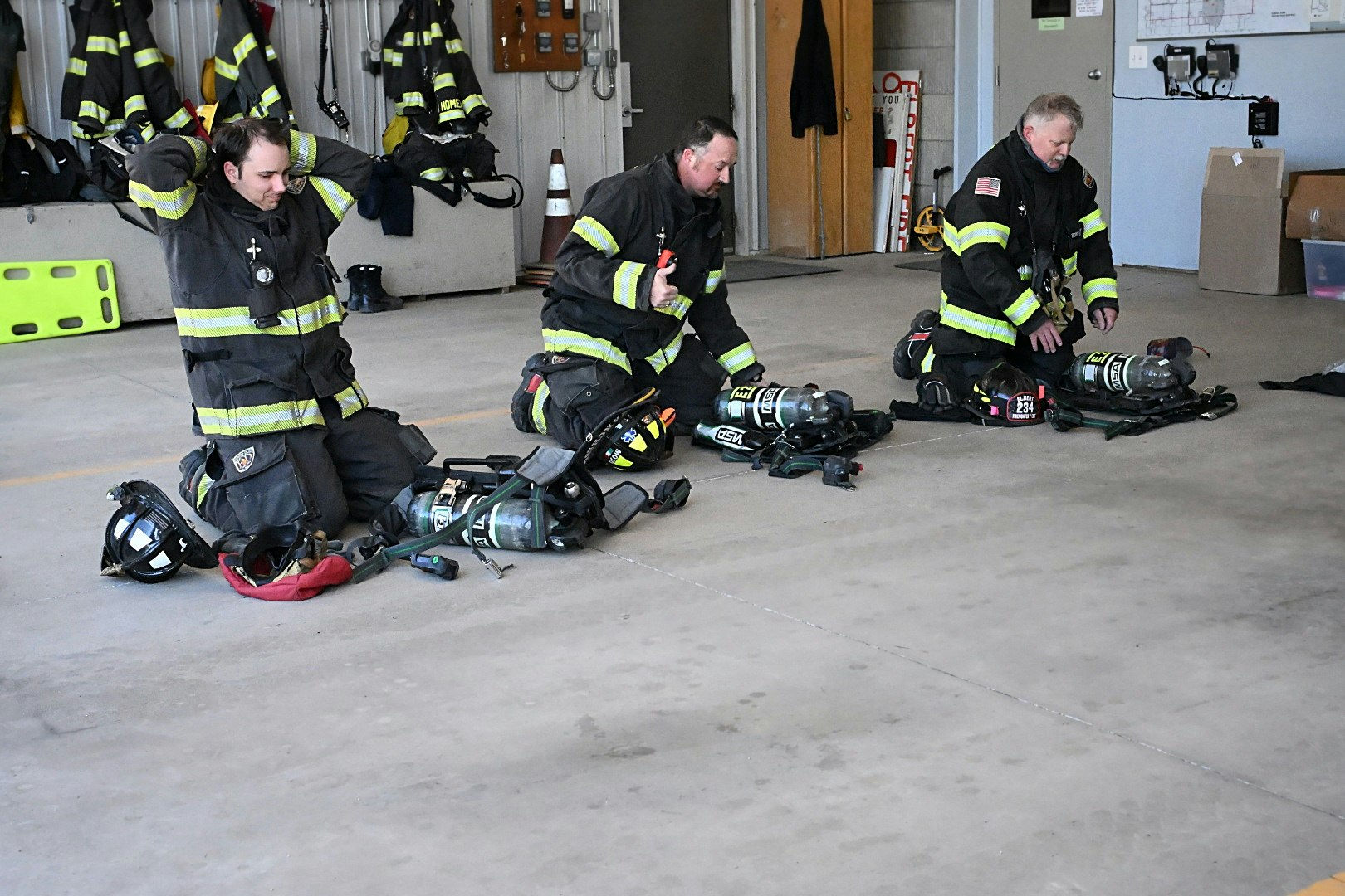 Three firefighters kneeling with gear inside a fire station, preparing equipment.