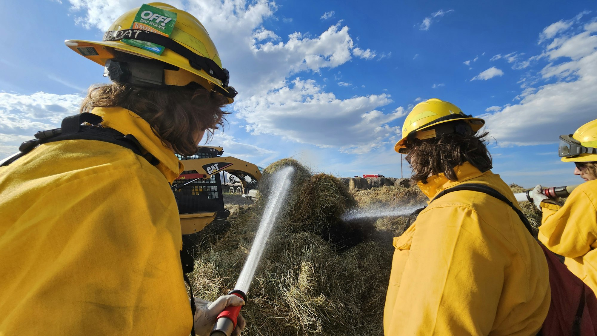 Firefighters in yellow helmets and gear spraying water on hay. Construction machinery and a clear blue sky in the background.