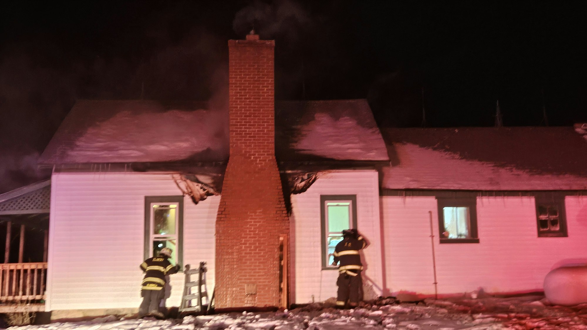 Firefighters outside a house with a brick chimney, roof damage, and smoke.