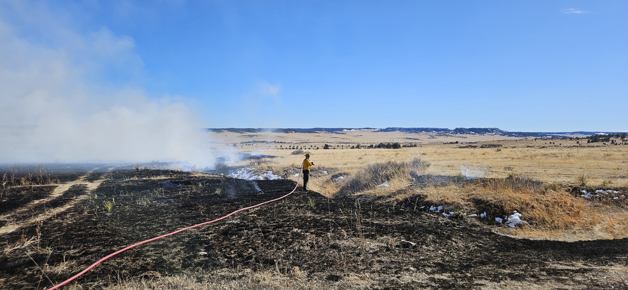 A firefighter is managing a grassland fire with a hose, surrounded by burnt and unburnt grass, with hills in the background.