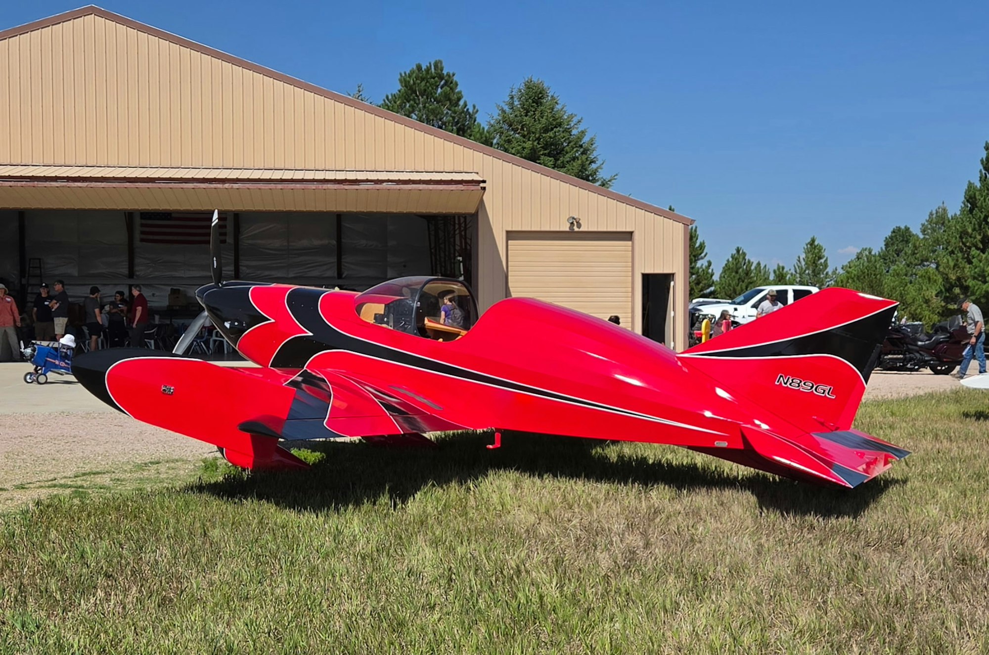 A striking red and black aircraft sits on grass outside a hangar, with people visible in the background.