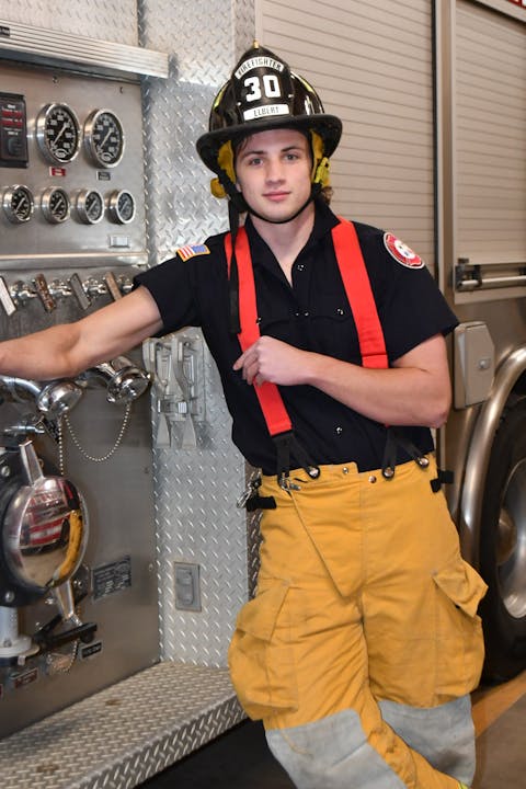 A firefighter in uniform and helmet leans against a fire truck, next to equipment controls.