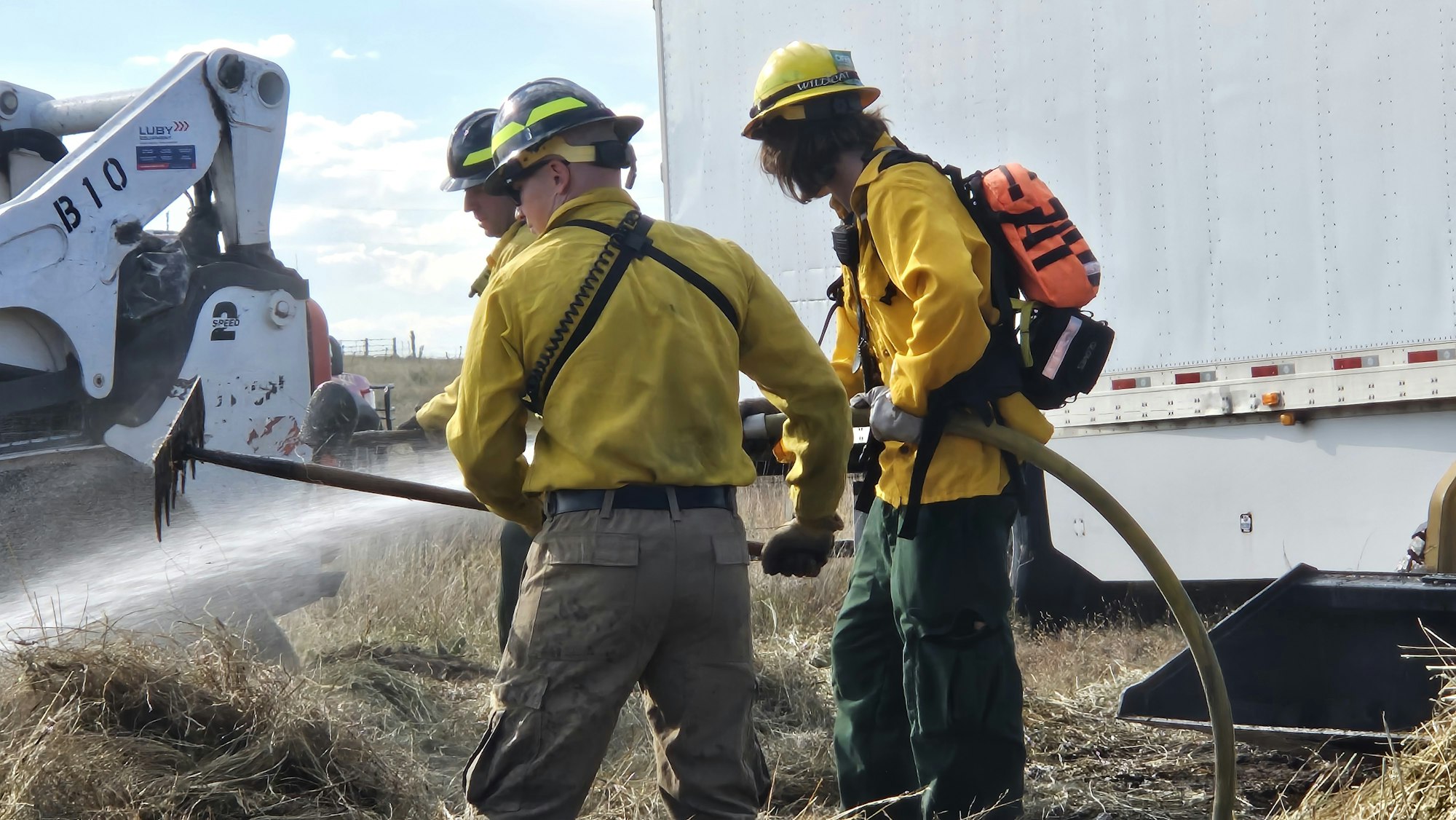 Two firefighters in yellow gear and helmets next to a white truck, handling a hose spraying water.