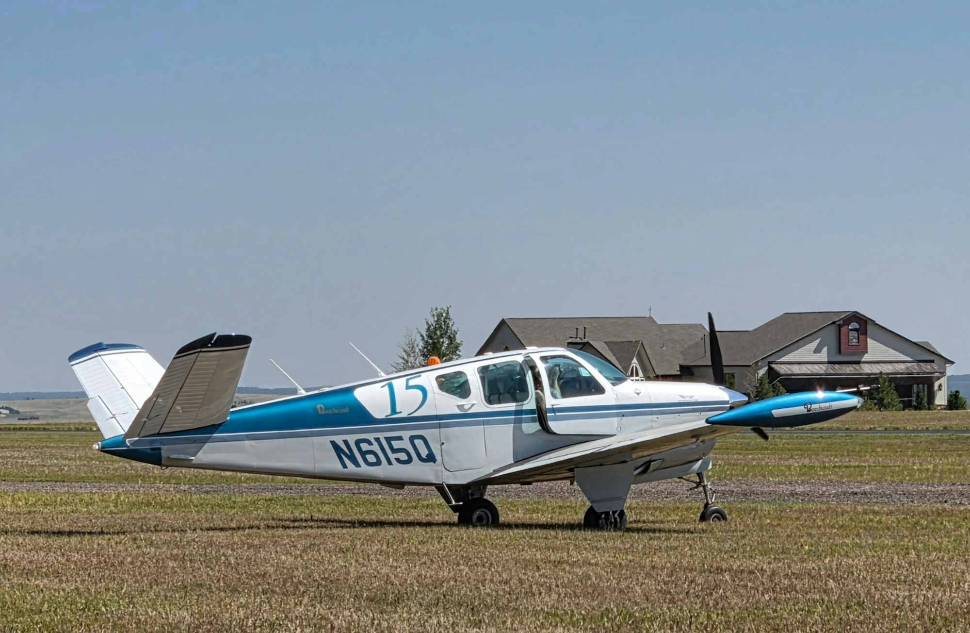 A single-engine airplane is parked on grass, with a residential house visible in the background under a clear blue sky.