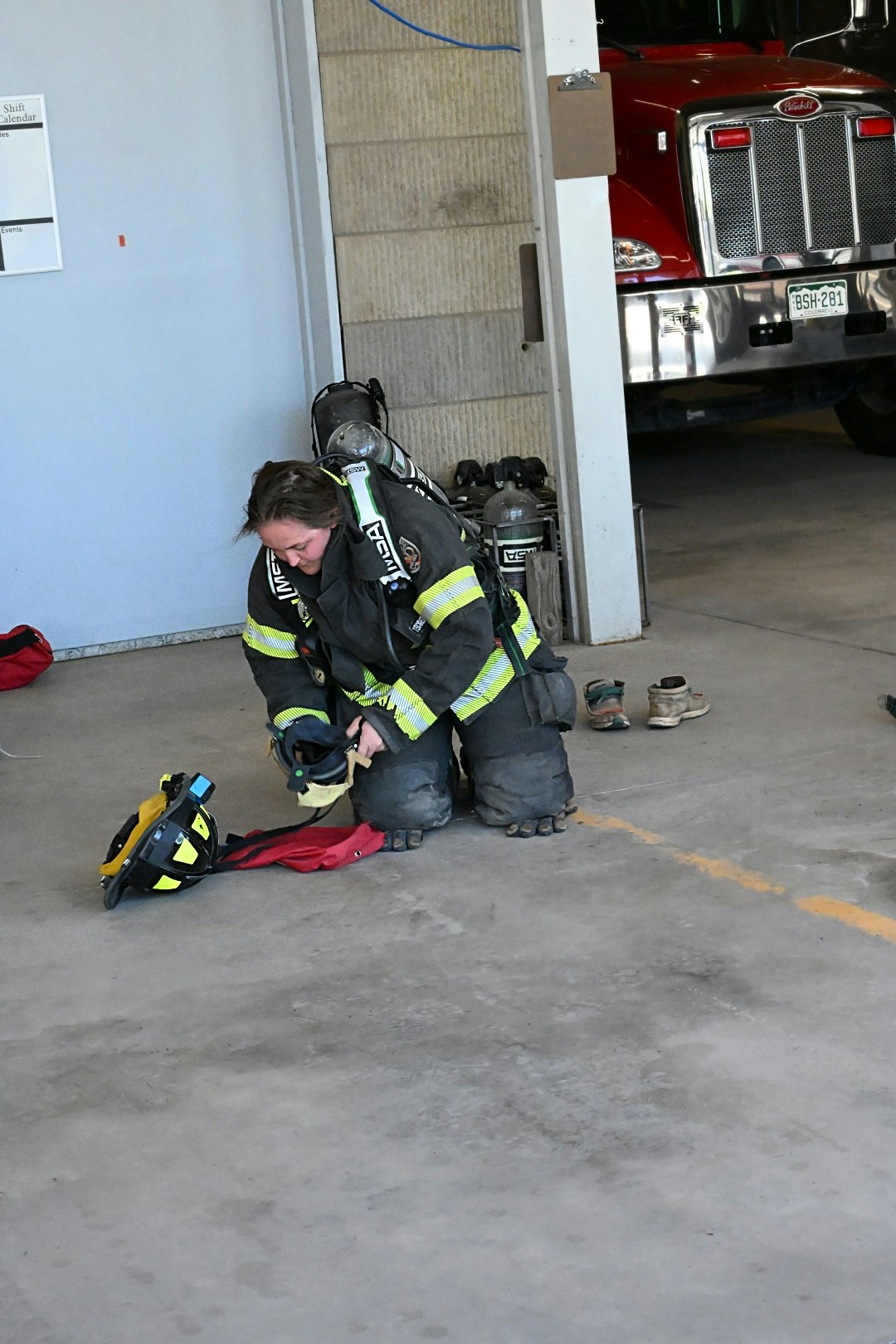 A firefighter kneels, organizing gear in a fire station near a red fire truck.