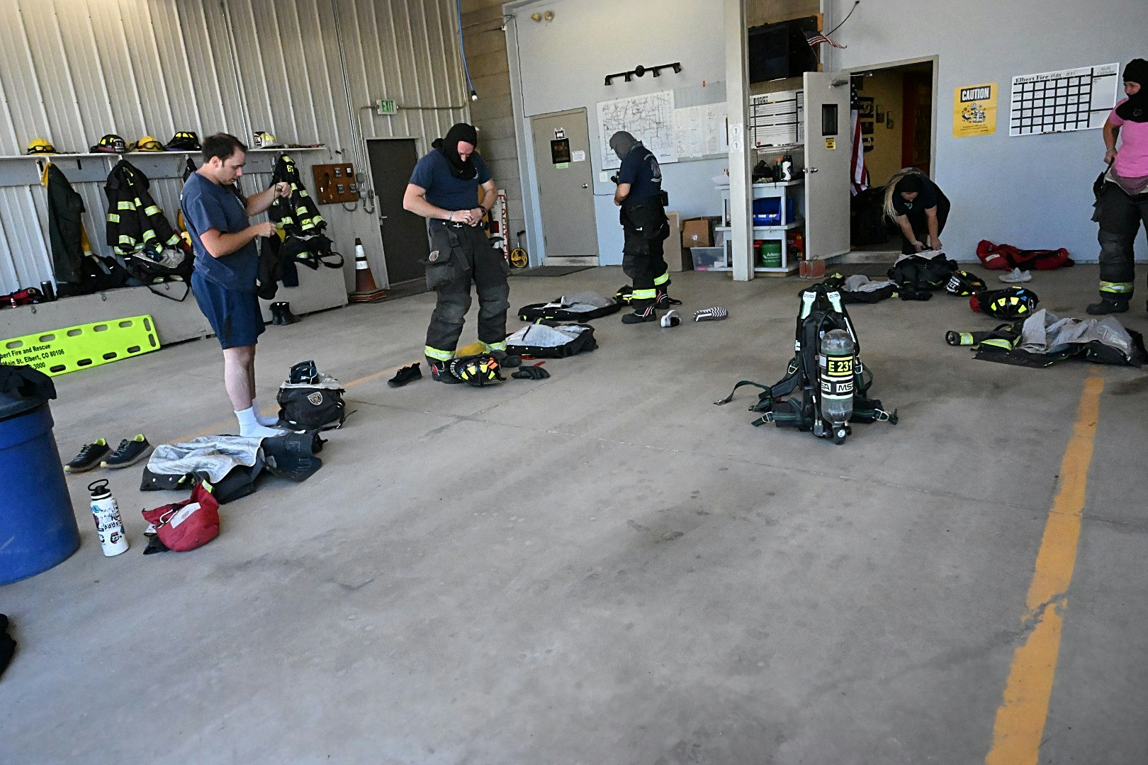 Firefighters donning gear in a garage with equipment and helmets on shelves.