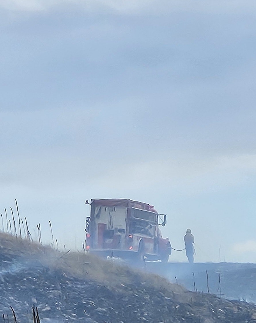 A fire truck is parked on a hill as a firefighter uses a hose, with smoke and burned grass surrounding the area.