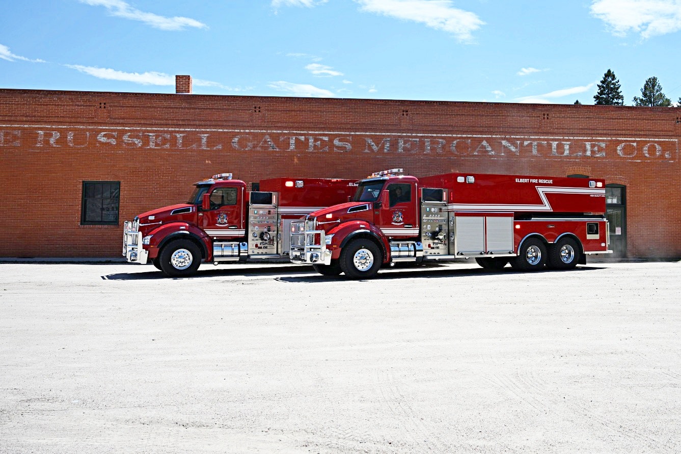 Two red fire trucks are parked in front of a brick building with faded signage, under a clear blue sky.
