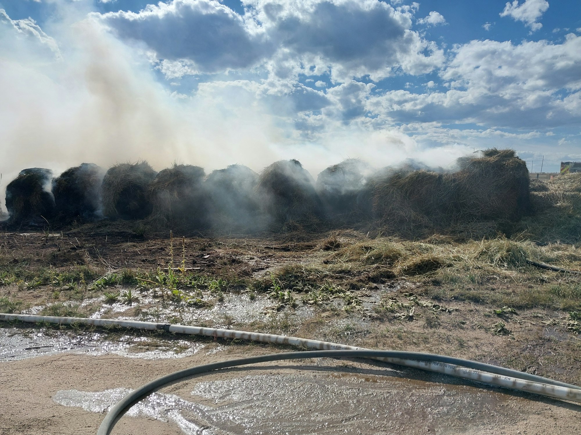 Hay bales with smoke, blue sky with clouds, and a muddy foreground.