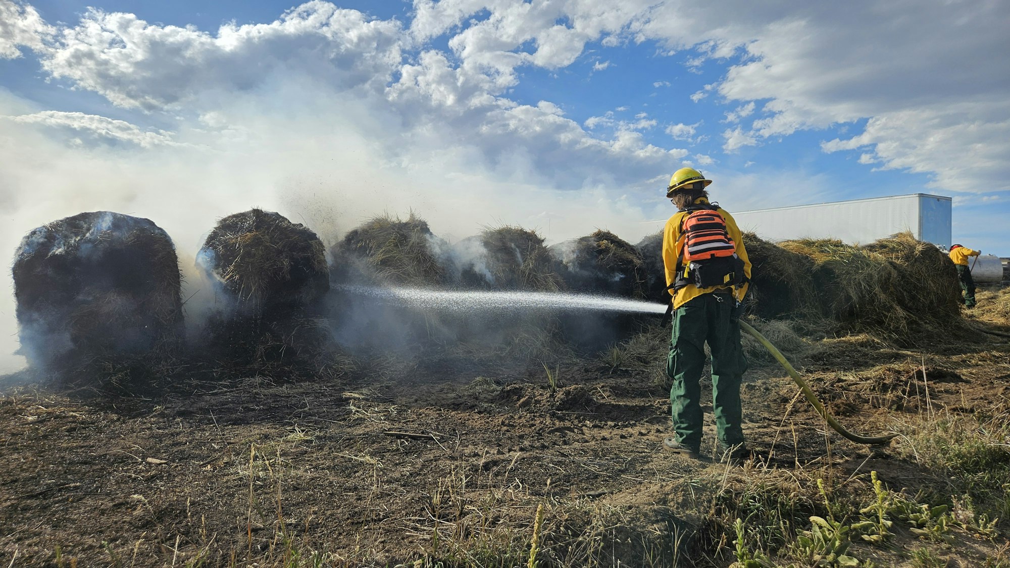 Firefighters extinguishing smoldering hay bales under a cloudy sky.