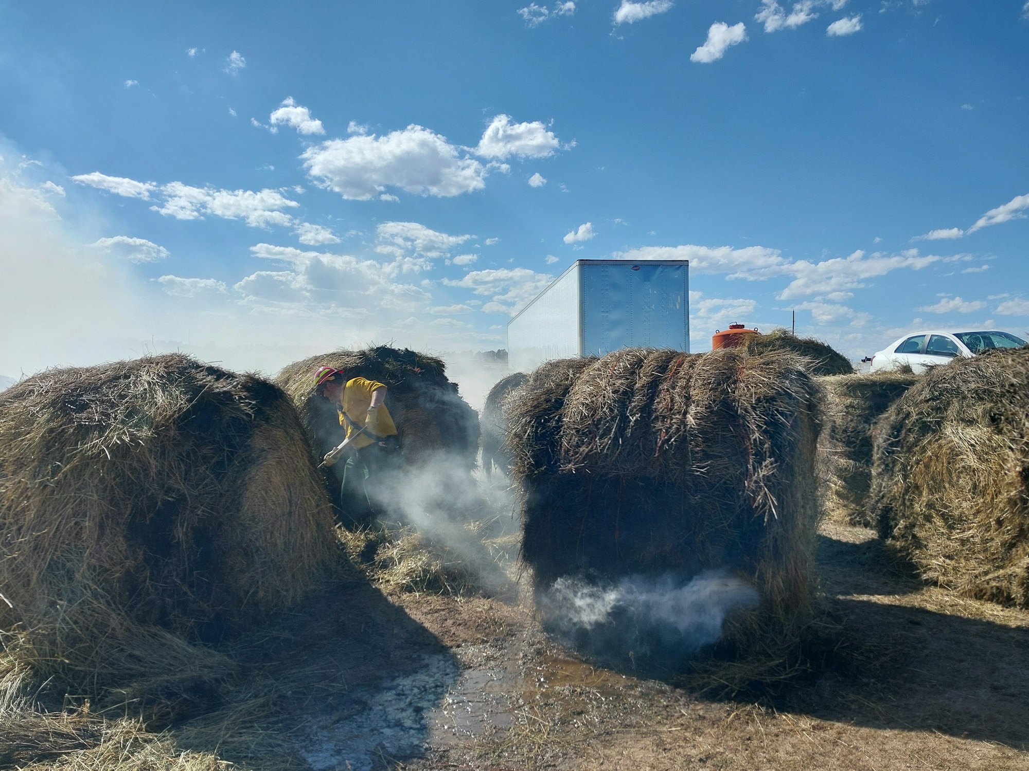 Person extinguishing a hay bale fire under a blue sky with clouds.