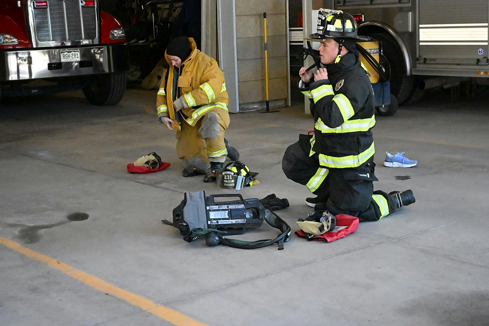 Two firefighters kneeling, getting into their gear in a fire station with trucks and equipment nearby.