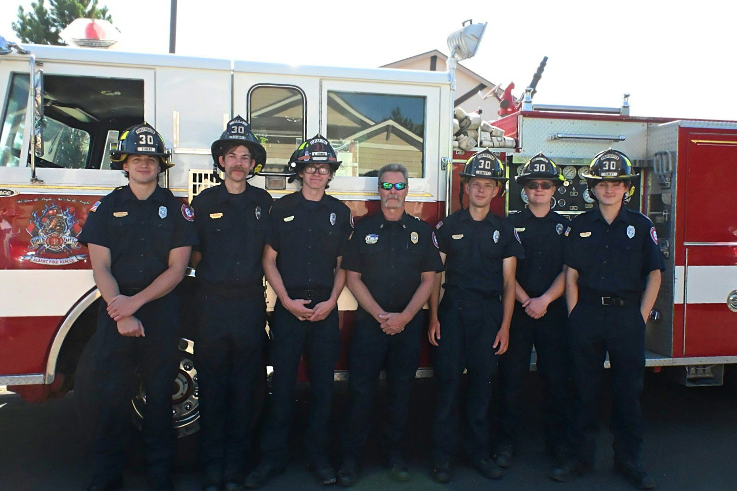 A group of seven firefighters in uniform standing in front of a fire truck.