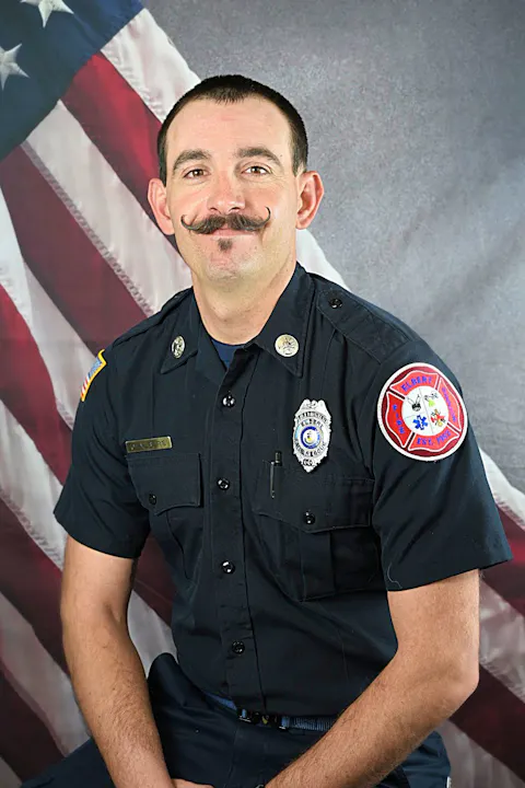 A man in a fire department uniform poses in front of a backdrop featuring the American flag, showcasing a distinctive mustache.