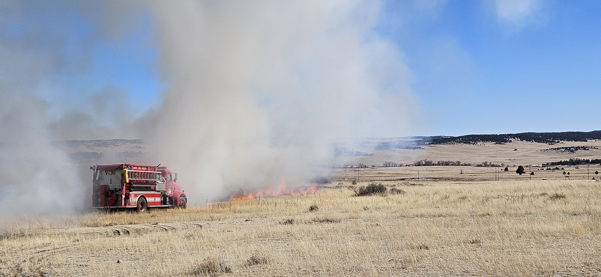 A fire truck battles a grass fire in a large open field with smoke clouding the sky.
