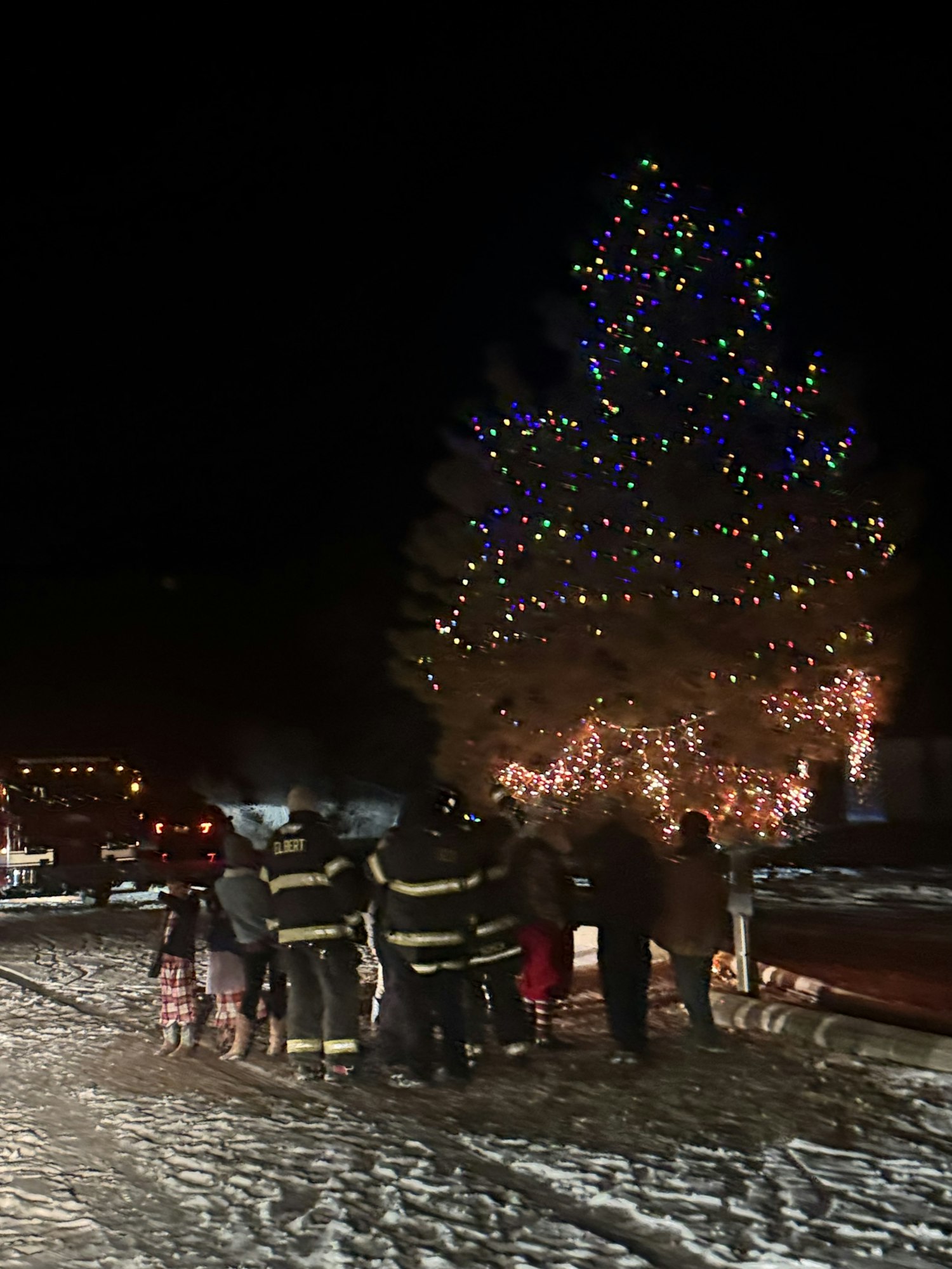 People in winter clothing gather near a large, decorated Christmas tree at night.