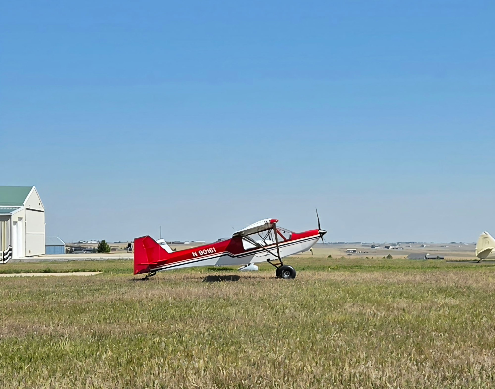A red small aircraft is parked on a grassy area near a building, with a clear blue sky in the background.