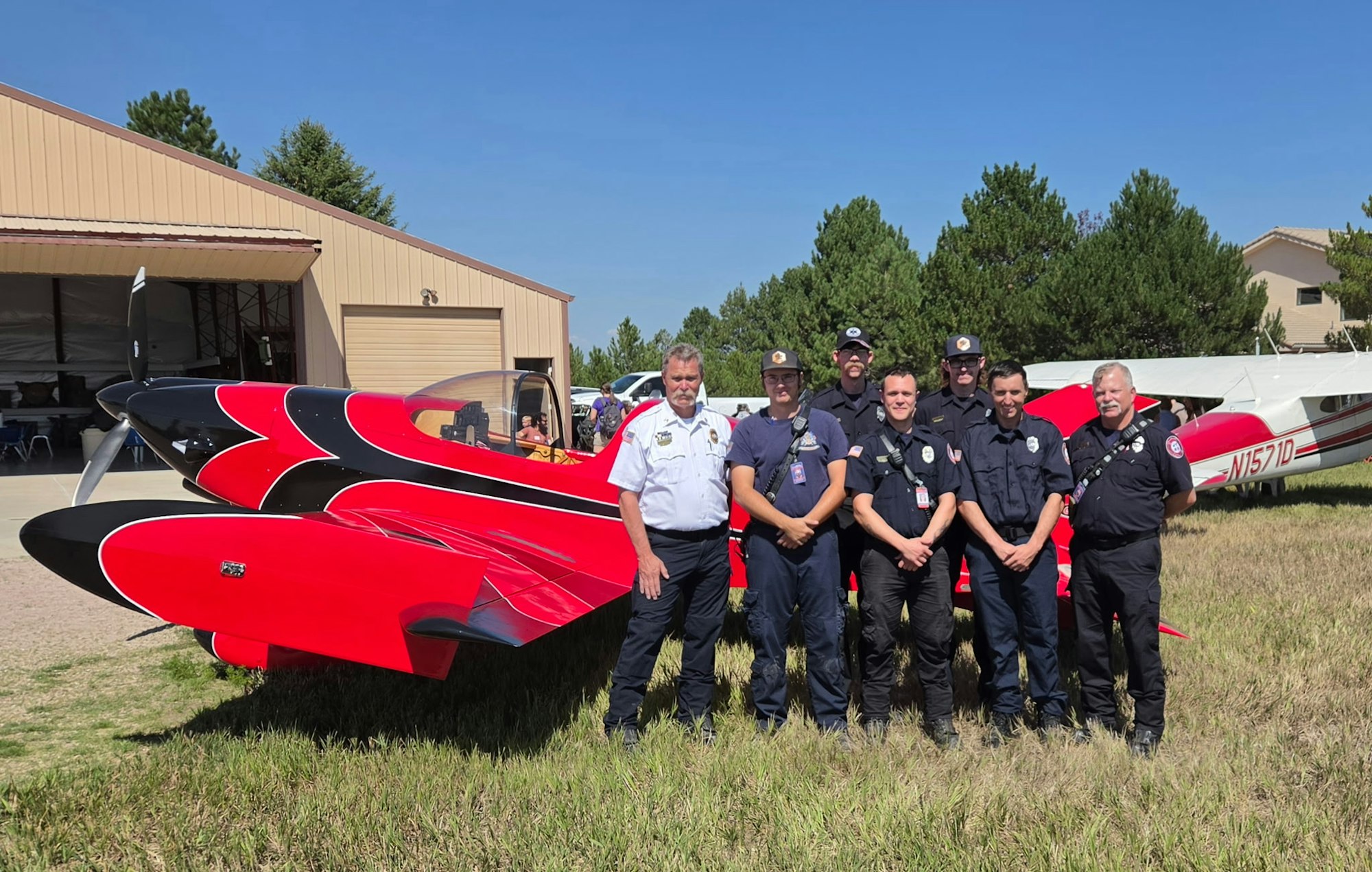 A group of emergency personnel poses in front of a vibrant red aircraft and a hangar on a sunny day.