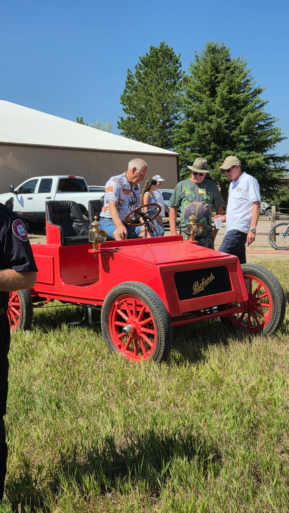 A group of people examines a vintage red Packard car in a grassy area with trees and buildings in the background.