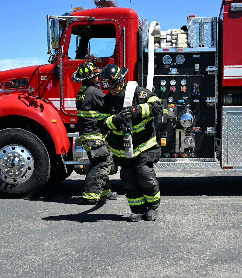 Firefighters in full gear are managing a fire hose beside a red fire truck.