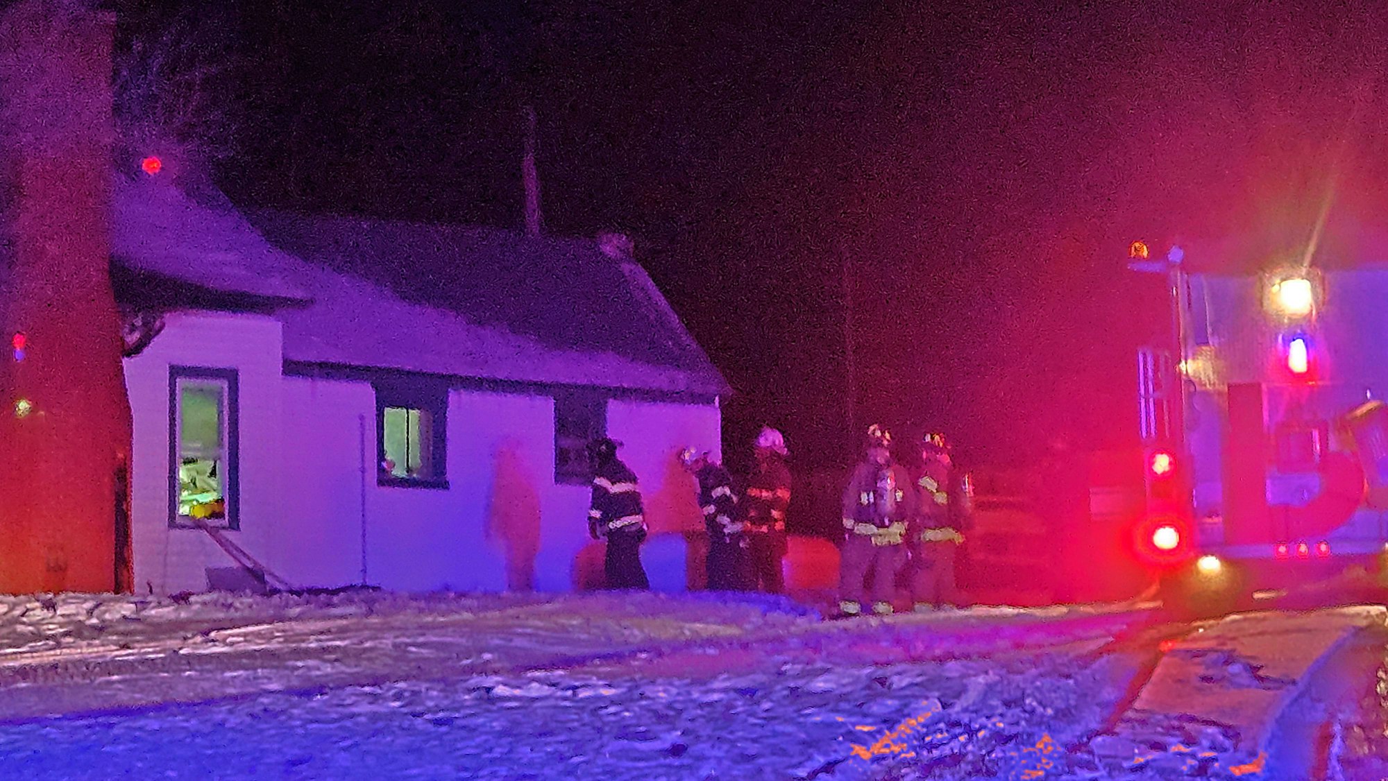 Firefighters in gear beside a house at night, illuminated by emergency vehicle lights.