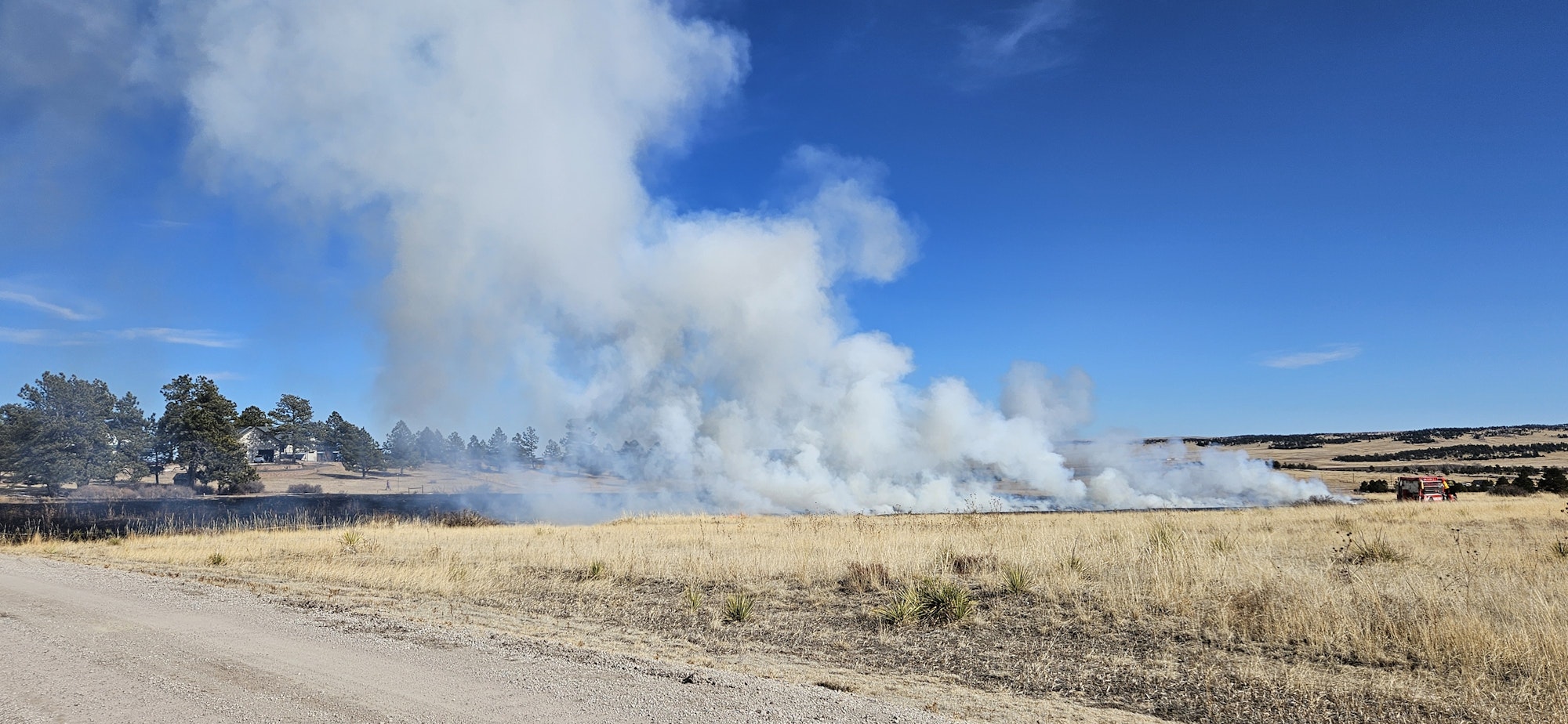 A controlled burn with smoke on grassland, clear sky, and a fire truck on the right.