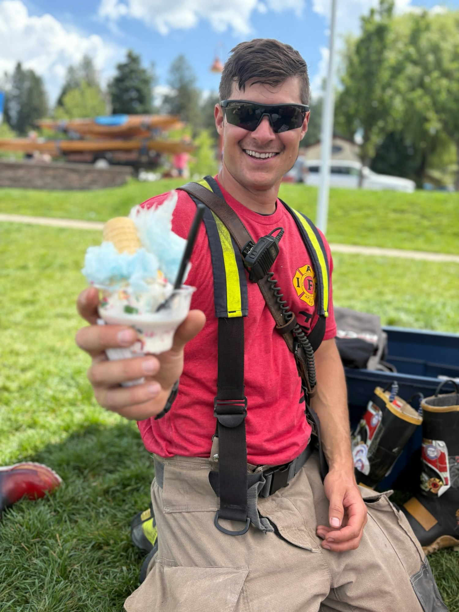 A firefighter in a red shirt enjoys ice cream topped with cotton candy, smiling on a grassy field with kayaks in the background.