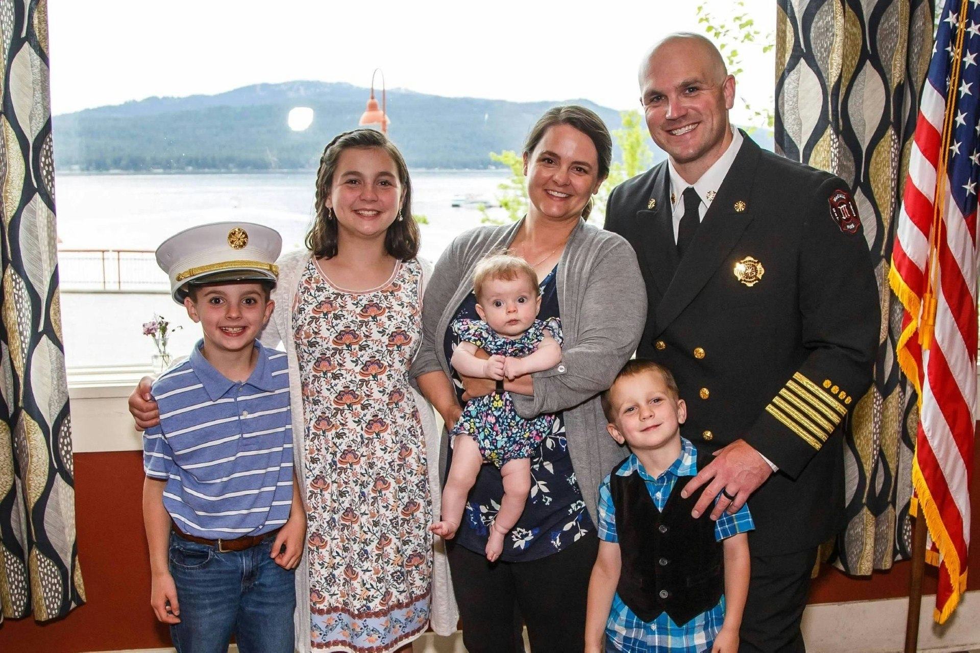 A family of six poses indoors; a man in uniform, a woman, and four children. There's an American flag and a lake view.