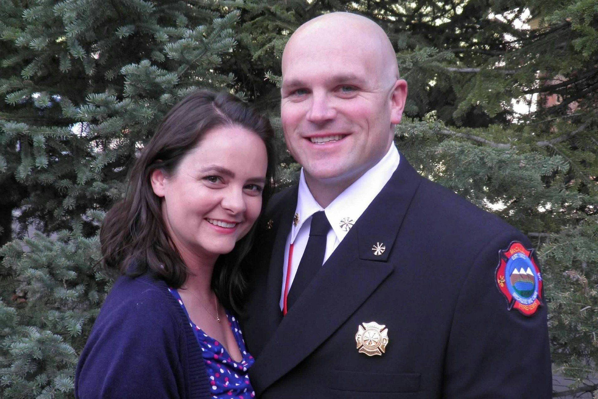 A smiling couple, one in a blue uniform with fire department insignia, pose in front of green pine trees.