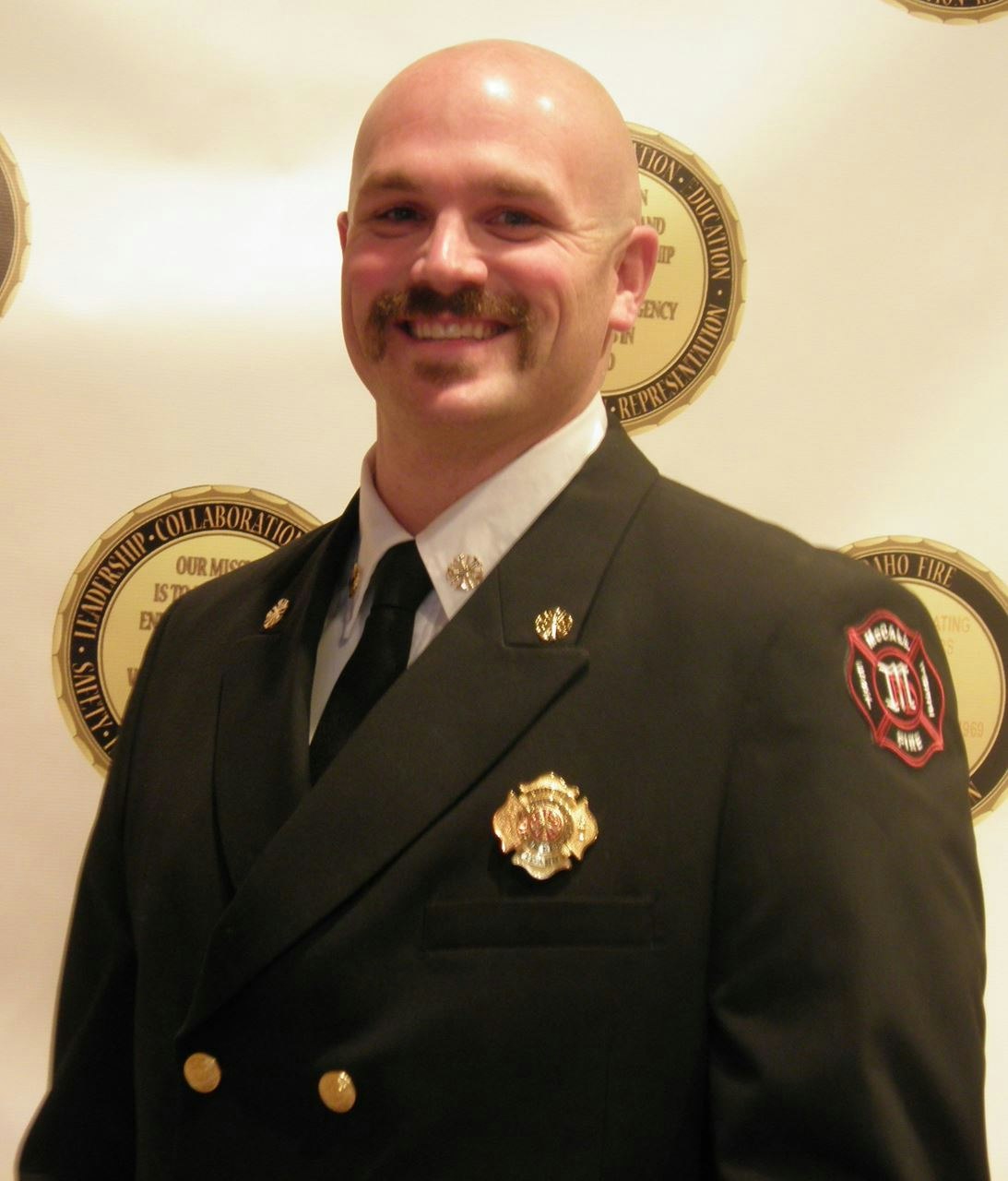 A smiling person in a firefighter's dress uniform with badges, in front of a backdrop with emblems.