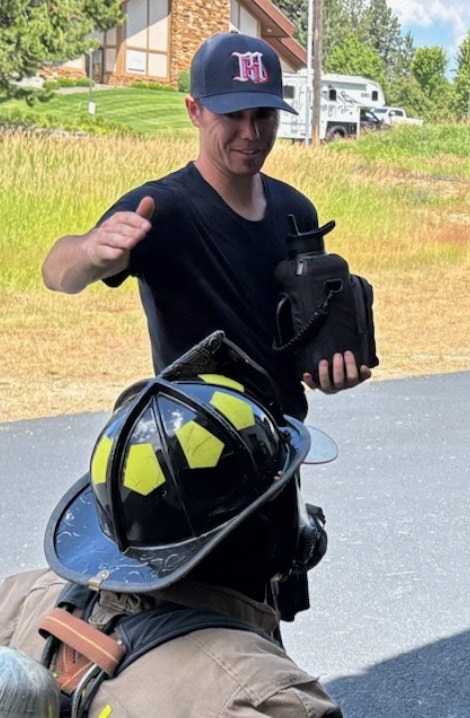 A person handing something to a firefighter outside, with a house and greenery in the background. The firefighter's helmet has yellow stickers.