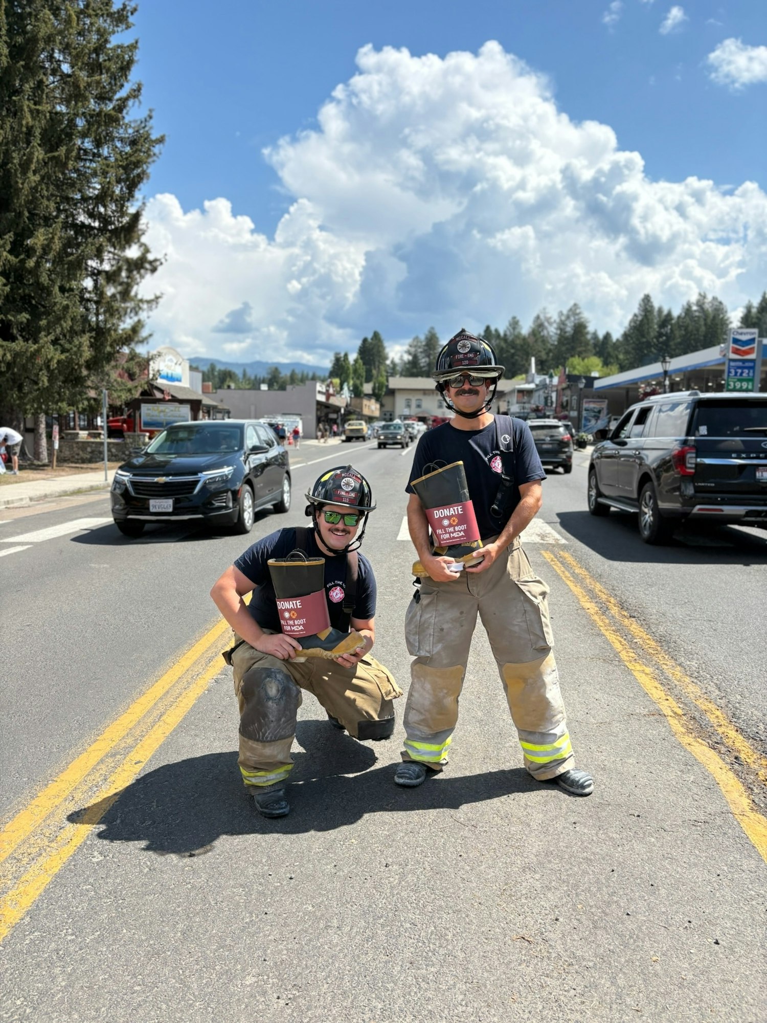 Two firefighters in gear are asking for donations in the middle of a busy street on a sunny day.